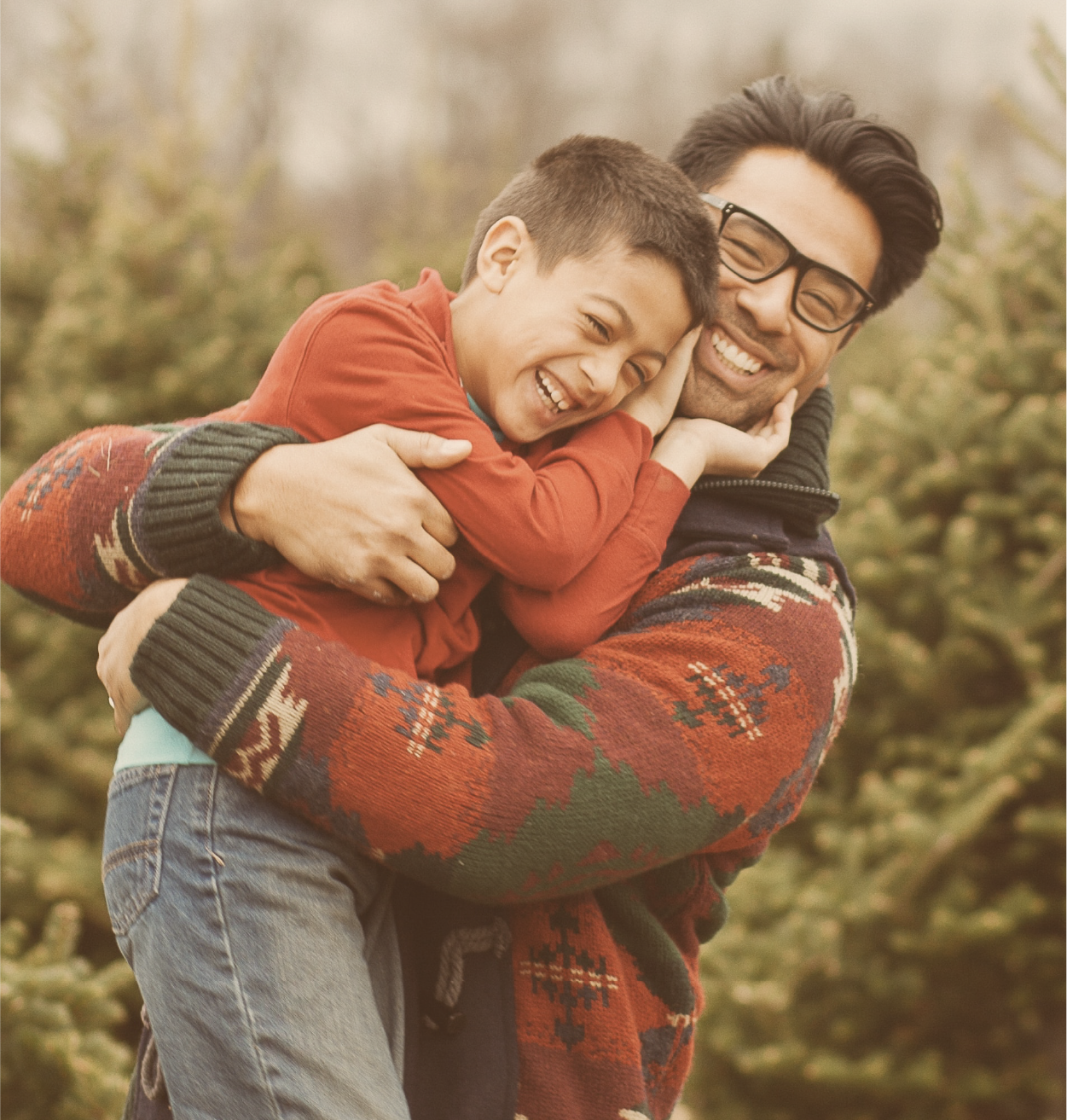 Père souriant portant son fils dans ses bras devant des sapins